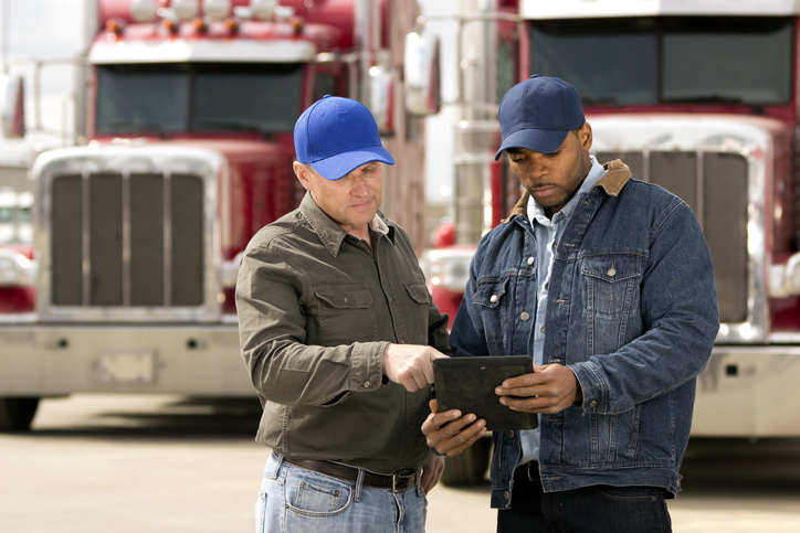 Two men speaking while looking at a tablet with two semi-trucks behind them.